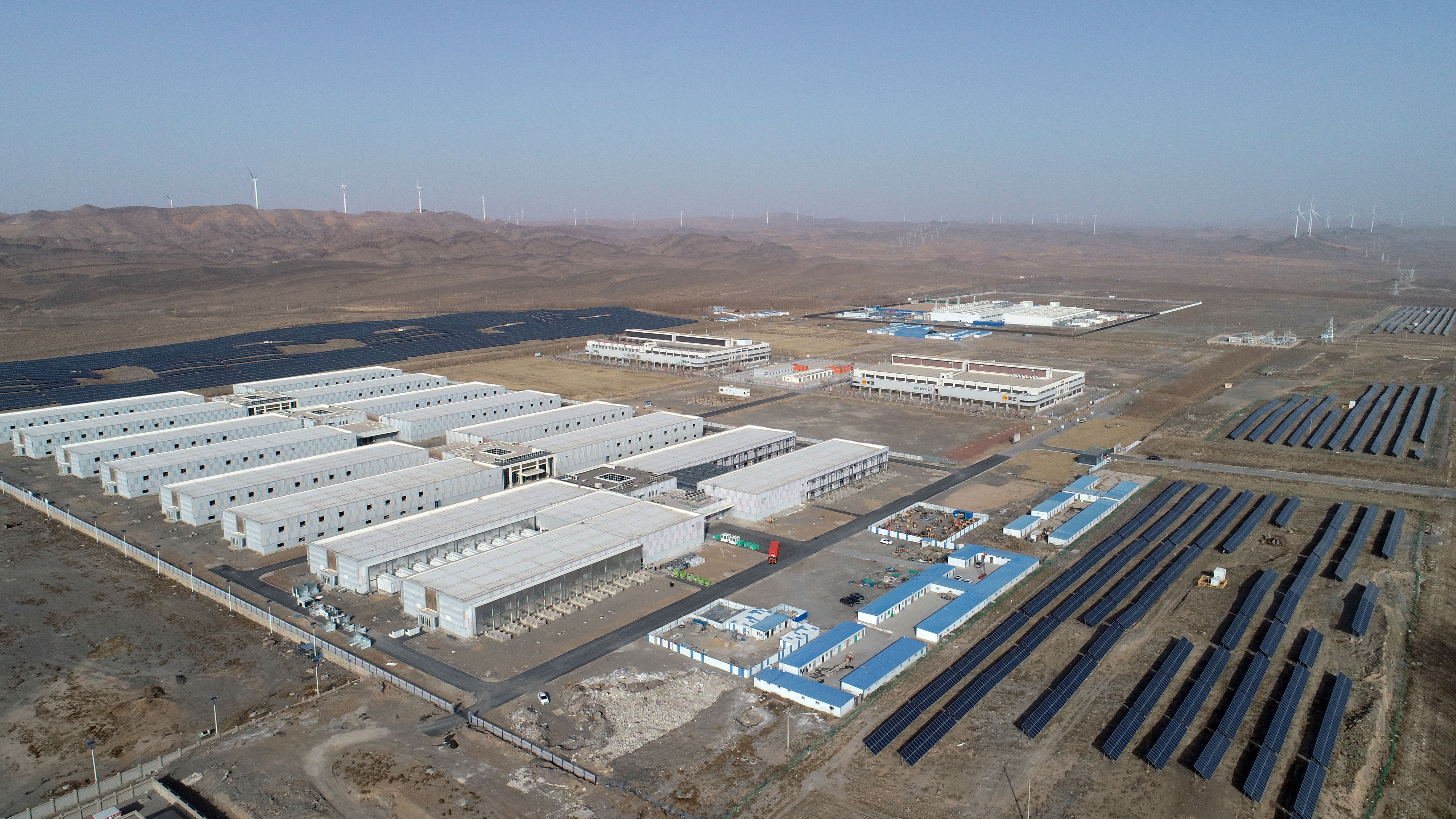 A sprawling data center located in a desert with wind turbines in the background