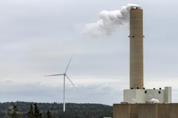 A wind turbine generator behind the smokestack of a fossil fuel generating plant. Overcast sky.