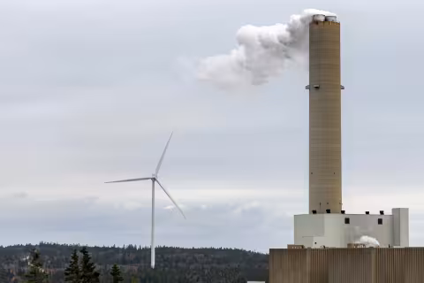 A wind turbine generator behind the smokestack of a fossil fuel generating plant. Overcast sky.